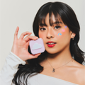 Model wearing pastel pink, blue, and purple acne patches. Purple heart with yellow sparkle, Pink Cat Head Character, Blue Clouds with sparkle and moon. Model is holding a pastel pink rounded square compact that contains acne patches.