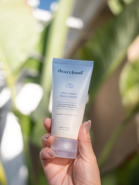 A hand holding a yellow blue gradient moisturizer tube of 3.7 fl oz. Ultra-hydrating moisturizer in front of a blurry plant. 