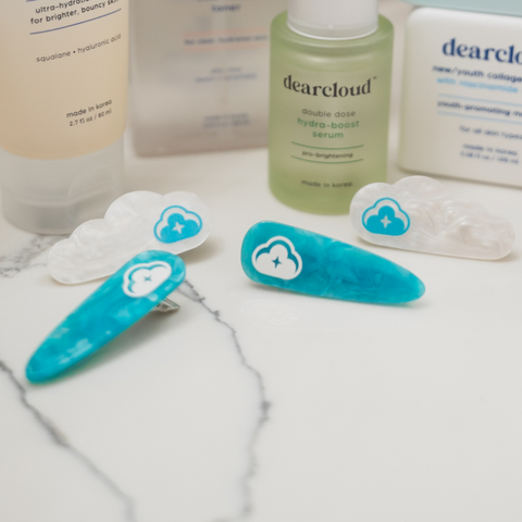 Four hair clips. First pair of shiny flecked blue hair pins with white dearcloud cloud image. Second pair of shiny flecked white cloud shaped hair pinks with same blue dearcloud cloud image. Both on a bathroom counter in front of other skincare products. 