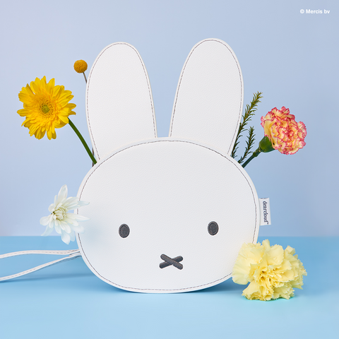 A white bag with a Miffy bunny face design is adorned with yellow and pink flowers placed in and around the ears. The background is light blue.